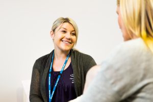 An over the shoulder image of a blonde female Mercy Health worker smiles at a woman to her left.