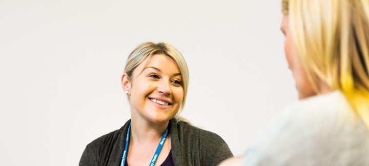 An over the shoulder image of a blonde female Mercy Health worker smiles at a woman to her left.