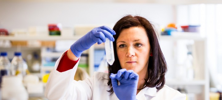 A medium close up of a woman in a lab wearing blue gloves and white lab coat holding a test tube.